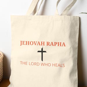 May include: A cream-colored canvas tote bag featuring the text JEHOVAH RAPHA in orange, a black cross graphic, and the text THE LORD WHO HEALS in orange. The bag has two long fabric handles and is constructed from a plain, light-colored textile material.