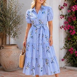 Könnte beinhalten: Ein blau-weiß gestreiftes Midikleid mit vertikalen Streifen, Kragen, kurzen Puffärmeln, durchgehender Knopfleiste und einem passenden Stoffgürtel in der Taille. Das Kleid ist mit einem floralen Muster in Blau, Grün und Rosa verziert.