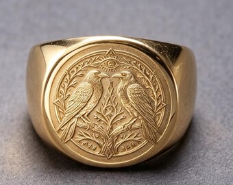May include: A gold-toned signet ring featuring a circular face with an embossed design of two birds perched on a central tree branch. Above the birds is an eye symbol, and the entire scene is framed by a decorative border with intricate leaf patterns.