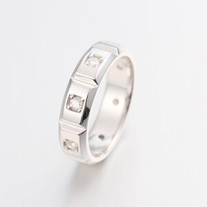 May include: A polished silver-toned metal band ring featuring a series of three square-set clear round stones embedded along the outer surface. The ring has a smooth, reflective finish with a structured design that highlights the three distinct settings.