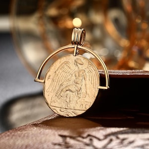 Könnte beinhalten: Goldfarbener Anhänger mit detailliertem Münzdesign, das eine gravierte geflügelte Figur zeigt. Die Münze wird in einem kreisförmigen Rahmen gehalten, der an einer Öse mit mehreren Schlaufen befestigt ist. Der Anhänger ist aus Metall und für Halsketten geeignet.