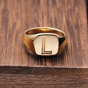 Könnte beinhalten: Ein goldfarbener Siegelring mit einer flachen, quadratischen Oberfläche, in deren Mitte der Großbuchstabe L eingraviert ist. Der Ring hat eine polierte, reflektierende Oberfläche und eine glatte, abgerundete Schiene, die nach hinten schmaler wird.