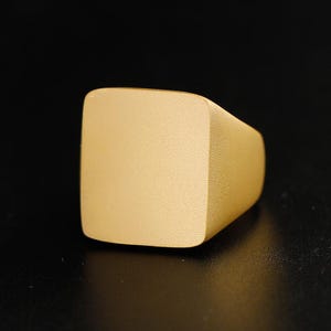 Può includere: Un anello a sigillo color oro con una faccia quadrata. L'anello ha una finitura opaca e un design semplice e moderno. L'anello è posto su uno sfondo nero.
