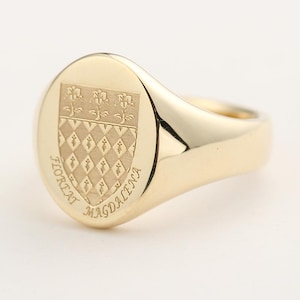 May include: A gold signet ring with an oval face. The face features a detailed crest with floral and diamond patterns, and the inscription "FLOREAT MAGDALENA" is etched around the crest. The ring has a polished finish.