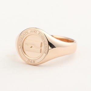 May include: A rose gold signet ring with a circular emblem. The emblem features the words "LANDSCAPE AND ENGINEERING" around the edge and the letters "D&B" at the bottom. The center of the emblem has a stylized "Z" shape.