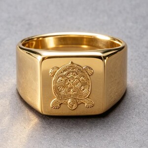 Peut inclure: Une bague chevalière en métal doré poli avec un motif de tortue gravé en détail sur la face carrée plate. L'anneau est large et lisse avec une finition métallique réfléchissante, présentant une représentation claire et centrée d'une tortue.