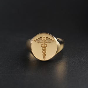 Puede incluir: Anillo de sello dorado con una cara circular. La cara presenta un grabado detallado del bastón de Asclepio, un símbolo de la medicina. El anillo tiene un acabado pulido y un diseño clásico.