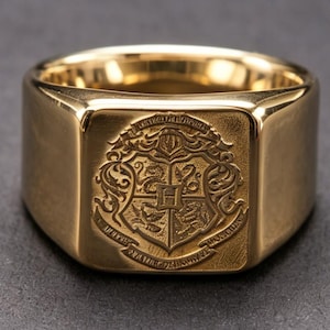 May include: A gold-colored signet ring with a rectangular face. The face of the ring features an embossed crest with intricate details. The ring has a polished finish and a classic, bold design.