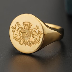 May include: A gold signet ring with a detailed crest. The crest features two lions flanking a shield with the letter "H" inside. The ring has a polished finish and a classic, rounded design. A timeless piece of jewelry.