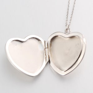 May include: A silver-toned heart-shaped locket pendant suspended from a delicate matching chain. The locket is shown in an open position, revealing two smooth, concave interior surfaces. The metal has a polished finish with a bright, reflective sheen.