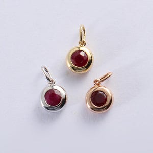 May include: Three gemstone pendants. One pendant is gold, one is silver, and one is rose gold. Each pendant features a round, faceted, deep red ruby gemstone set in a bezel setting. The pendants have a small loop for a chain.
