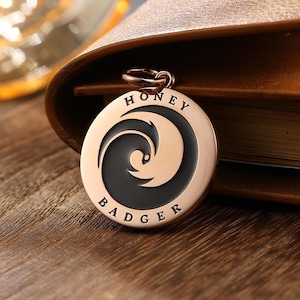 Op de afbeelding: Een ronde, roségouden hondenpenning met de woorden "HONEY BADGER" rond de rand gegraveerd. De penning heeft een zwart, gestileerd das-ontwerp in het midden. De penning is bevestigd aan een roségouden ring.
