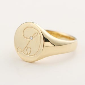 Op de afbeelding: Een gouden zegelring met een gepolijste afwerking. De ring heeft een verhoogde, cirkelvormige voorkant met de letter "L" gegraveerd in een scriptlettertype en een kleine diamantaccent. De band is glad en afgerond.