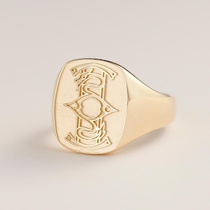 May include: A gold signet ring with a rectangular face featuring an engraved design. The design includes a central eye-shaped element, flanked by stylized serpent-like figures. The ring has a polished finish.
