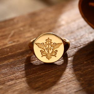 Op de afbeelding: Een gouden zegelring met een gegraveerd wapenontwerp. Het wapen heeft een kroon, vleugels en sierlijke details. De ring ligt op een houten oppervlak.