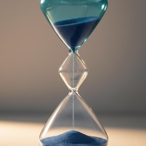 May include: An elegant hourglass with a teal top and base, and a clear central section. The hourglass is filled with blue sand, with some sand flowing from the top to the bottom. The design is symmetrical, suitable for home decor or as a gift.