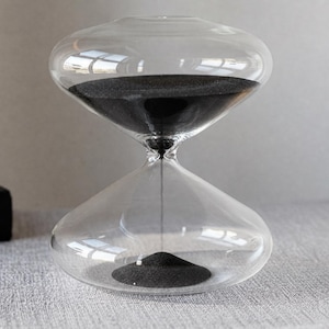 May include: A clear glass hourglass with black sand. The hourglass has a bulbous top and bottom, connected by a narrow neck. The black sand is partially in the bottom bulb, with the rest in the top, indicating time has passed.