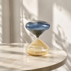 May include: A glass hourglass featuring a translucent blue top bulb and a translucent amber bottom bulb. The two sections are joined at a narrow centre point, creating a symmetrical shape with smooth, rounded curves and a polished finish.