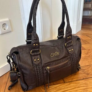 Può includere: Una borsa a mano marrone scuro con superficie testurizzata, dotata di due manici, una tasca frontale con cerniera e una targhetta con logo in metallo dorato con la scritta Flora. La borsa presenta dettagli laterali intrecciati e finiture in ottone.