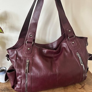 Könnte beinhalten: Eine tiefburgunderrote Schultertasche mit zwei vertikalen Reißverschlusstaschen auf der Vorderseite, zwei Griffen mit silberfarbenen Metallbeschlägen und seitlichen Einstecktaschen. Die Tasche hat eine glatte, strukturierte Oberfläche mit sichtbaren Nähten.