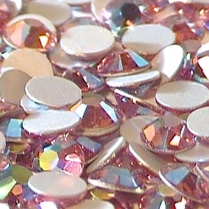 May include: Close-up of a collection of iridescent rhinestones. The rhinestones are a mix of shapes, including round and faceted, with a pink and iridescent color. The flat-backed rhinestones are scattered, creating a sparkling effect.