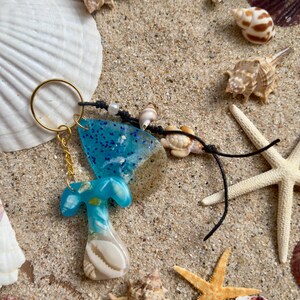 Könnte beinhalten: Ein handgefertigter Schlüsselanhänger mit goldenem Ring und blauem Harzdesign, mit einer Muschel und Glitzerakzenten. Der Schlüsselanhänger ist von Muscheln und Seesternen auf sandiger Oberfläche umgeben, was ein Strandthema hervorruft.