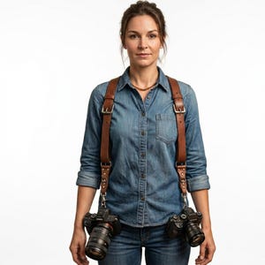 May include: A brown leather dual camera strap with metal buckles and hardware. Two black cameras are attached to the strap. The photographer is wearing a denim shirt and jeans.