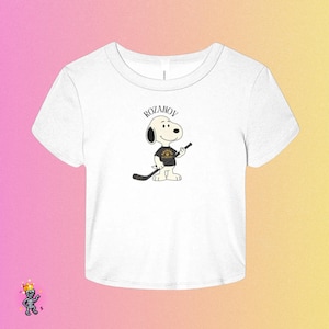 May include: White crop top with a cartoon Snoopy character holding a hockey stick. Snoopy is wearing a black shirt with a gold emblem and the text "ROZANOV" above. The top is a casual, short-sleeved style.