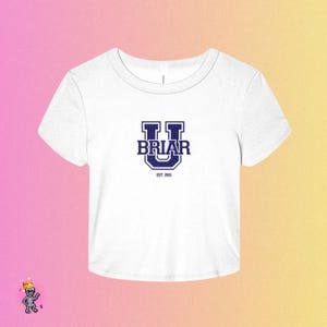 May include: A white short-sleeved crop top featuring a dark blue collegiate-style graphic on the chest. The design displays a large letter U with the word BRIAR centered inside it, and the text EST. 2015 printed in a smaller font directly below.