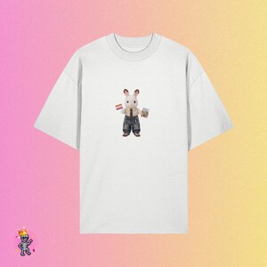 Puede incluir: Una camiseta blanca de manga corta con un gr&aacute;fico central de un peque&ntilde;o conejo con corbata marr&oacute;n y vaqueros azules. El conejo sostiene una peque&ntilde;a bandera a rayas naranja, blanca y rosa en una mano y un peque&ntilde;o libro rectangular en la otra.