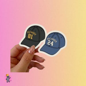 May include: Two baseball cap stickers. One is black with gold text reading "ROZANOV 81". The other is blue with white text reading "HOLLANDER 24". Both stickers are on a gradient background.