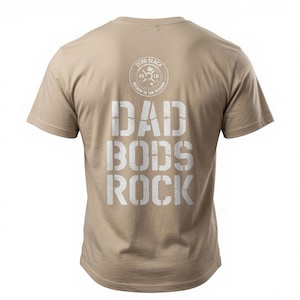 May include: A tan short-sleeved t-shirt featuring white distressed text on the back that reads DAD BODS ROCK. Above the text is a circular graphic resembling a weight plate with the words ZERO SLACK and ROCKIN IS THE REASON printed.