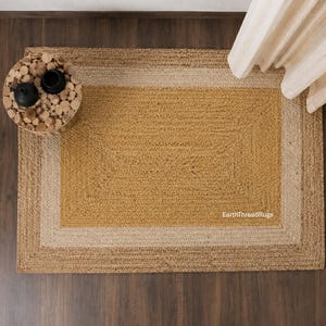 May include: A rectangular jute rug with a three-tone design. The center is a warm gold, framed by a light beige border, and a darker brown outer edge. The rug is on a dark wood floor.