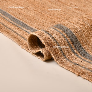 Op de afbeelding: Close-up van een opgerolde jute vloerkleed met grijze strepen. Het vloerkleed heeft een natuurlijke bruine kleur met een geweven textuur. De grijze strepen lopen horizontaal over het vloerkleed. Het vloerkleed is gedeeltelijk opgerold en toont de onderkant.