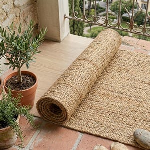 Könnte beinhalten: Eine aufgerollte Jute-Rolle in Naturtönen liegt auf einer Ziegelterrasse, teilweise ausgerollt, um ihre gewebte Textur zu zeigen. Ein kleiner Topf-Olivenbaum und andere Pflanzen sind im Vordergrund. Die Abmessungen des Teppichs sind nicht sichtbar.