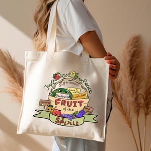 Può includere: Una borsa tote color crema con un'illustrazione colorata di frutta e le parole "Frutto dello Spirito". Il design include un'anguria, pomodoro, uva e altra frutta, con parole come "Gioia", "Pace" e "Bontà".