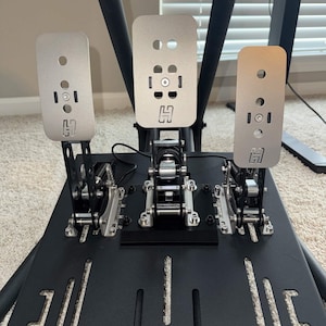 May include: A set of three silver-colored metal racing pedals mounted on a black base. Each pedal features a vertical rectangular plate with a small H logo, circular cutouts, and exposed mechanical springs and hinges for simulation gaming.