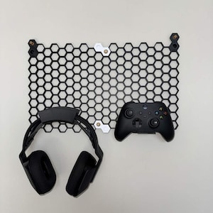 May include: Black gaming headset and controller displayed on a black honeycomb wall mount. The wall mount has a geometric design and is mounted on a white wall. The controller is black with multiple buttons.