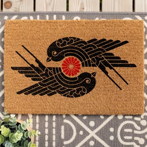 May include: A rectangular tan coir doormat featuring a black graphic print of two stylized birds facing each other in a circular formation. Between the birds is a small, vibrant red circular floral emblem with multiple radiating petals.