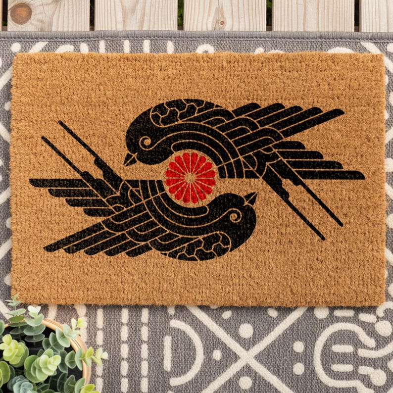 May include: A rectangular tan coir doormat featuring a black graphic print of two stylized birds facing each other in a circular formation. Between the birds is a small, vibrant red circular floral emblem with multiple radiating petals.