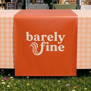 May include: A rectangular orange table runner featuring the white text "barely fine" in a stylized serif font. The text includes a decorative swirled graphic element integrated into the letter f, centered on the smooth, solid-colored fabric.