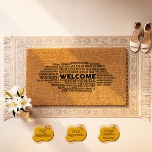 May include: A rectangular doormat with the word "WELCOME" in a variety of languages. The mat is a natural tan color with a decorative border. White heels and a vase of white flowers are next to the mat.