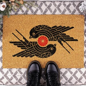 May include: A rectangular tan coir doormat featuring a black graphic print of two stylized birds facing each other in a circular formation, with a small red circular floral emblem positioned directly between the two bird figures.