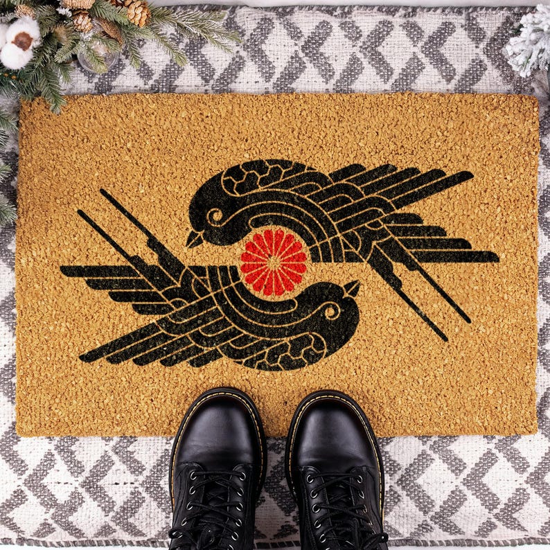 May include: A rectangular tan coir doormat featuring a black graphic print of two stylized birds facing each other in a circular formation, with a small red circular floral emblem positioned directly between the two bird figures.