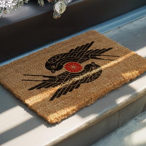 May include: A rectangular tan coir doormat featuring a black graphic print of two stylized birds facing opposite directions. Between the birds is a small, circular red floral emblem with multiple petals, creating a symmetrical design.