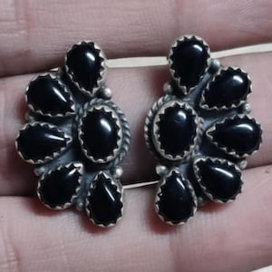 Puede incluir: Un par de pendientes de botón plateados, cada uno con un grupo de piedras negras en forma de lágrima. Las piedras están dispuestas en forma de flor, con un borde texturizado alrededor de cada grupo.