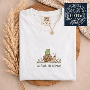 Puede incluir: Camiseta blanca Comfort Colors con cuello redondo, con un diseño bordado de un caracol que lleva una rana, con el texto "No Rush, No Worries". Un collar dorado con un colgante de sol está sobre la camiseta.