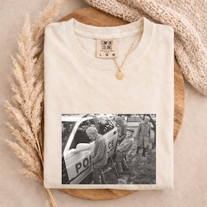 May include: Cream-colored Comfort Colors t-shirt with a black and white image of older men near a police car. The shirt has a small gold pendant necklace. The shirt is on a wooden surface.