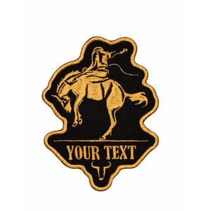May include: Embroidered patch featuring a cowboy riding a bucking bronco. The patch is black with gold-colored embroidery. The text "YOUR TEXT" is displayed below the image of the cowboy and horse, with a bull's head below the text.