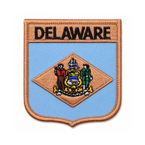 May include: Embroidered patch featuring the state of Delaware's emblem. The patch has a light blue background with a diamond-shaped emblem in the centre. The emblem includes two figures, a coat of arms, and the word "DELAWARE" at the top.